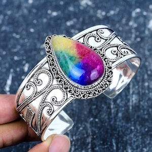 May include: A silver cuff bracelet with an elaborate design and a large, teardrop-shaped gemstone. The gemstone showcases a vibrant array of colours, including yellow, green, red, and purple. The bracelet's metalwork features intricate scroll patterns.
