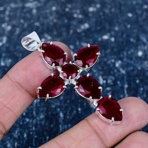 Puede incluir: Colgante de plata con forma de cruz, con cinco gemas facetadas de color rojo intenso en forma de lágrima. El colgante está sostenido por una mano sobre un fondo azul oscuro.