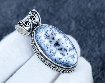 Puede incluir: Colgante ovalado con una piedra de ágata dendrítica, con un patrón de inclusiones blancas y negras. La piedra está engastada en un metal plateado con detalles ornamentados en los bordes y una fianza decorativa.