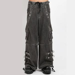 Puede incluir: Pantalones cargo de pierna ancha en mezclilla gris carbón con múltiples bolsillos utilitarios, anillas en D de metal plateado, correas ajustables con hebillas metálicas y tachuelas decorativas en las piernas. Incluyen cintura con cordón y botas de plataforma negras.