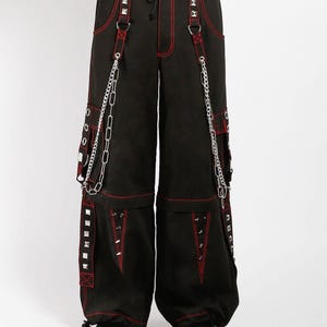 May include: Black pants with red stitching, featuring silver studs, chains, and straps. The pants have multiple pockets and a drawstring waist. The style is reminiscent of alternative fashion.