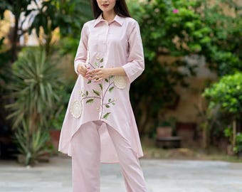 May include: A light pink two-piece outfit featuring a long-sleeved tunic top with floral embroidery and matching cropped pants. The tunic has a high-low hemline and a collared neckline. The outfit is worn with clear heels. The text "Paliwal_fashion" is visible.