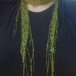 Puede incluir: Una bufanda larga de color verde oliva con una delicada textura de ganchillo de punto abierto, adornada con numerosas lentejuelas pequeñas y brillantes. La bufanda se envuelve alrededor del cuello, con dos extremos largos y finos que cuelgan.