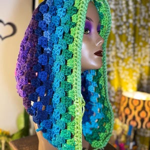 Op de afbeelding: Een handgemaakte gehaakte capuchon in een levendige kleurovergang van paars, blauw en groen. De capuchon wordt getoond op een mannequinhoofd en toont het getextureerde, open geweven ontwerp. De capuchon is een uniek accessoire.