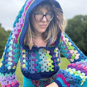 Könnte beinhalten: Ein farbenfrohes, gehäkeltes Kapuzentop mit Granny-Square-Design. Das Oberteil zeigt eine lebendige Mischung aus Blau-, Lila-, Grün- und Rosatönen. Die Kapuze und die Manschetten sind mit einem dunkleren Blau eingefasst, was einen auffälligen Kontrast erzeugt. Die Ärmel sind lang und ausgestellt.