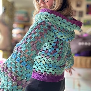 May include: A handmade crochet hoodie in shades of teal, blue, and purple. The jacket features a granny square pattern, a hood, and a purple trim along the hood and bottom hem. The sleeves are long and wide.