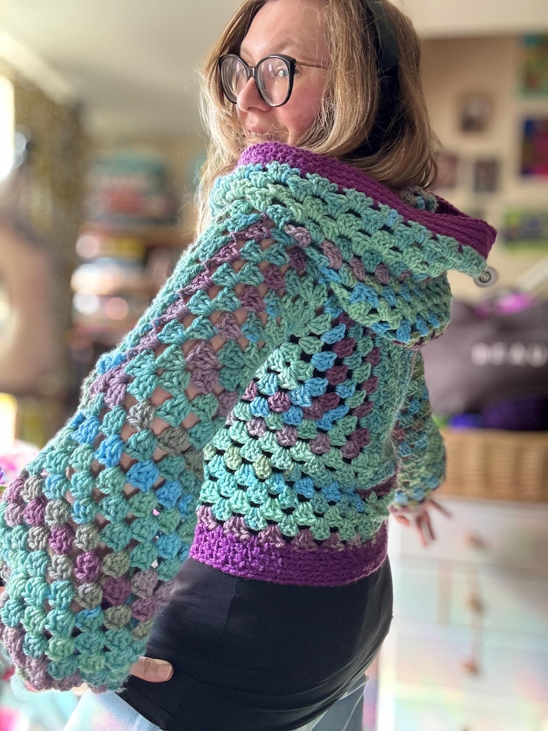 May include: A handmade crochet hoodie in shades of teal, blue, and purple. The jacket features a granny square pattern, a hood, and a purple trim along the hood and bottom hem. The sleeves are long and wide.