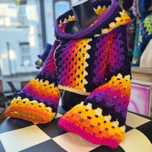 May include: A colorful, handmade crochet hooded top. The top features a vibrant gradient of colors, including purple, pink, orange, and yellow. The sleeves are flared, and the hood is trimmed in purple. The top is displayed on a black mannequin.