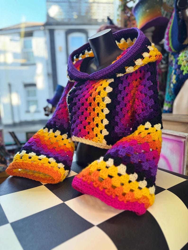 May include: A colorful, handmade crochet hooded top. The top features a vibrant gradient of colors, including purple, pink, orange, and yellow. The sleeves are flared, and the hood is trimmed in purple. The top is displayed on a black mannequin.