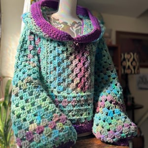 May include: A handmade crochet sweater in shades of green, blue, purple, and pink. The sweater features a large hood and wide sleeves with ribbed cuffs. The open weave design is visible throughout the garment.
