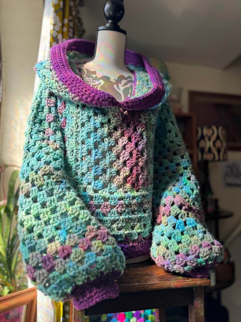 May include: A handmade crochet sweater in shades of green, blue, purple, and pink. The sweater features a large hood and wide sleeves with ribbed cuffs. The open weave design is visible throughout the garment.
