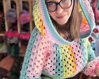 May include: A colorful, crocheted hooded sweater in a granny square pattern. The sweater features a rainbow of pastel colors, including pink, yellow, green, and blue. The sleeves have a flared design. The hood is lined with a contrasting color.