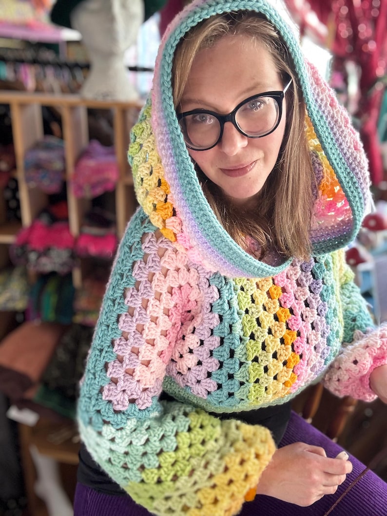 May include: A colorful, crocheted hooded sweater in a granny square pattern. The sweater features a rainbow of pastel colors, including pink, yellow, green, and blue. The sleeves have a flared design. The hood is lined with a contrasting color.