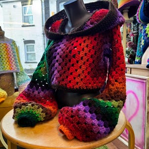 May include: A colorful, crocheted hooded jacket displayed on a mannequin. The jacket features a vibrant gradient of colors, including red, orange, purple, green, and brown. The sleeves and hood are prominently displayed, showcasing the intricate crochet pattern. The jacket is a unique, handmade garment.