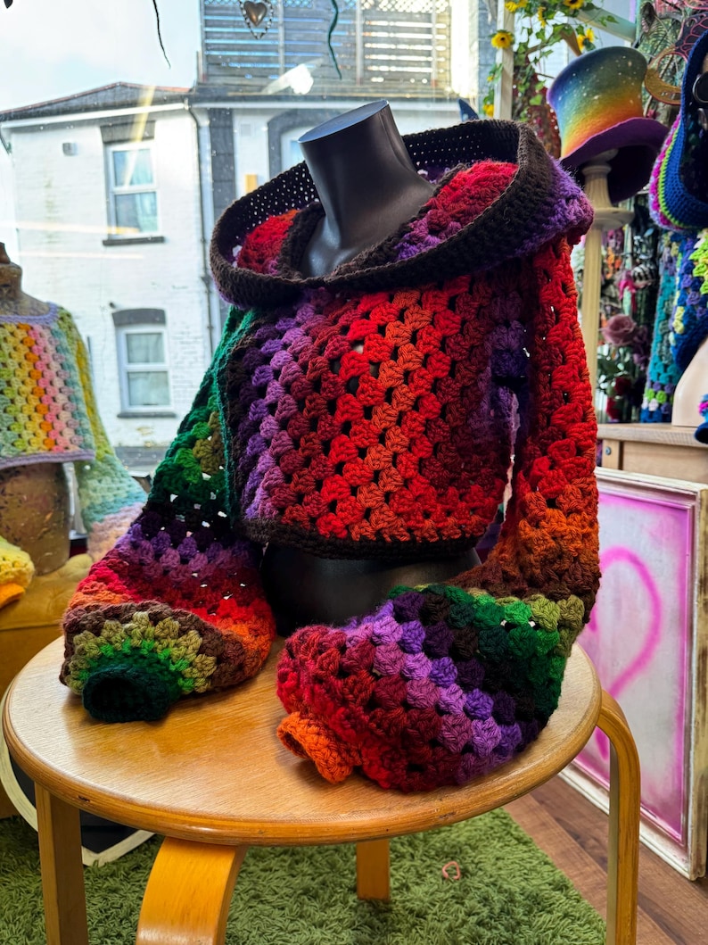 May include: A colorful, crocheted hooded jacket displayed on a mannequin. The jacket features a vibrant gradient of colors, including red, orange, purple, green, and brown. The sleeves and hood are prominently displayed, showcasing the intricate crochet pattern. The jacket is a unique, handmade garment.