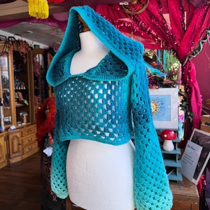 May include: A blue and teal crochet hooded shrug displayed on a mannequin. The shrug features a hood, long sleeves, and an openwork design. The color transitions from a deep blue to a lighter teal, creating an ombre effect. The garment is displayed on a white mannequin.