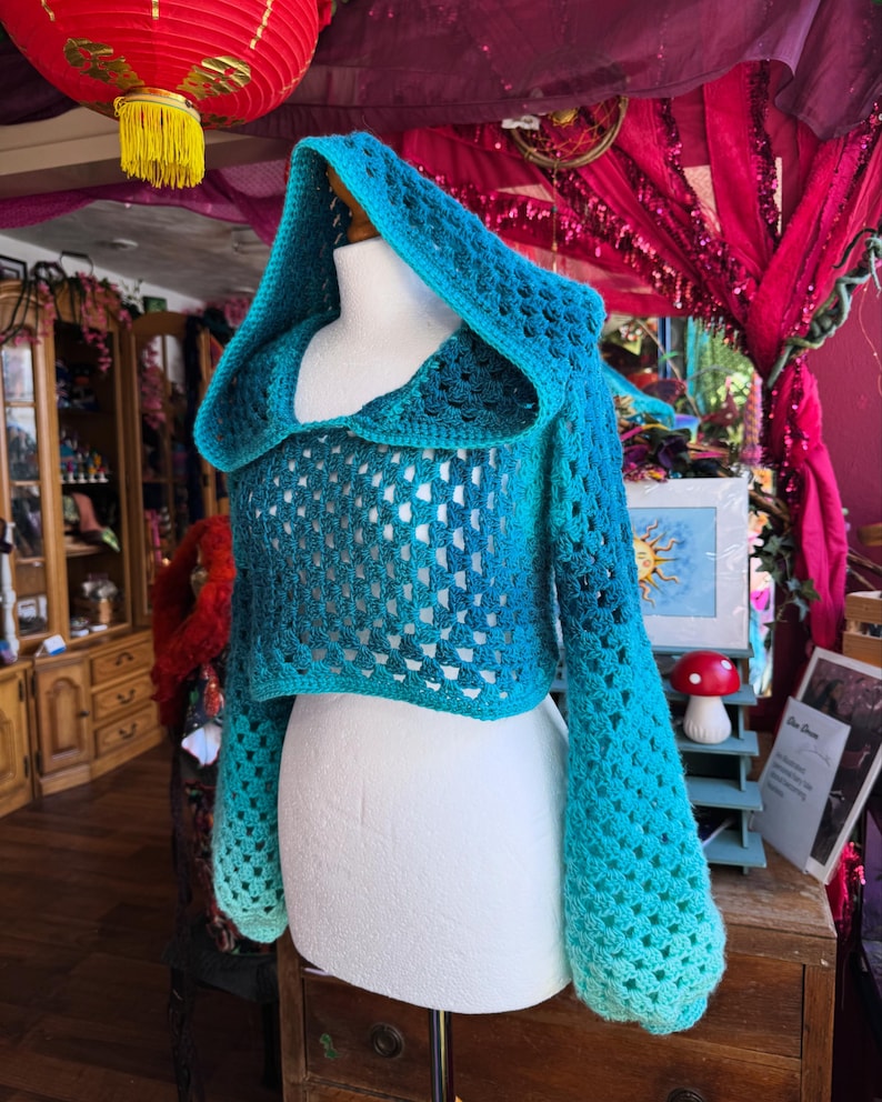 May include: A blue and teal crochet hooded shrug displayed on a mannequin. The shrug features a hood, long sleeves, and an openwork design. The color transitions from a deep blue to a lighter teal, creating an ombre effect. The garment is displayed on a white mannequin.