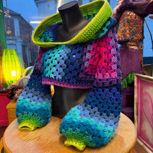 May include: A colorful, crocheted cropped sweater with a hood. The sweater features a vibrant gradient of blues, purples, and pinks, with a bright green hood and cuffs. The sleeves are long and puffy. The sweater is displayed on a black mannequin.