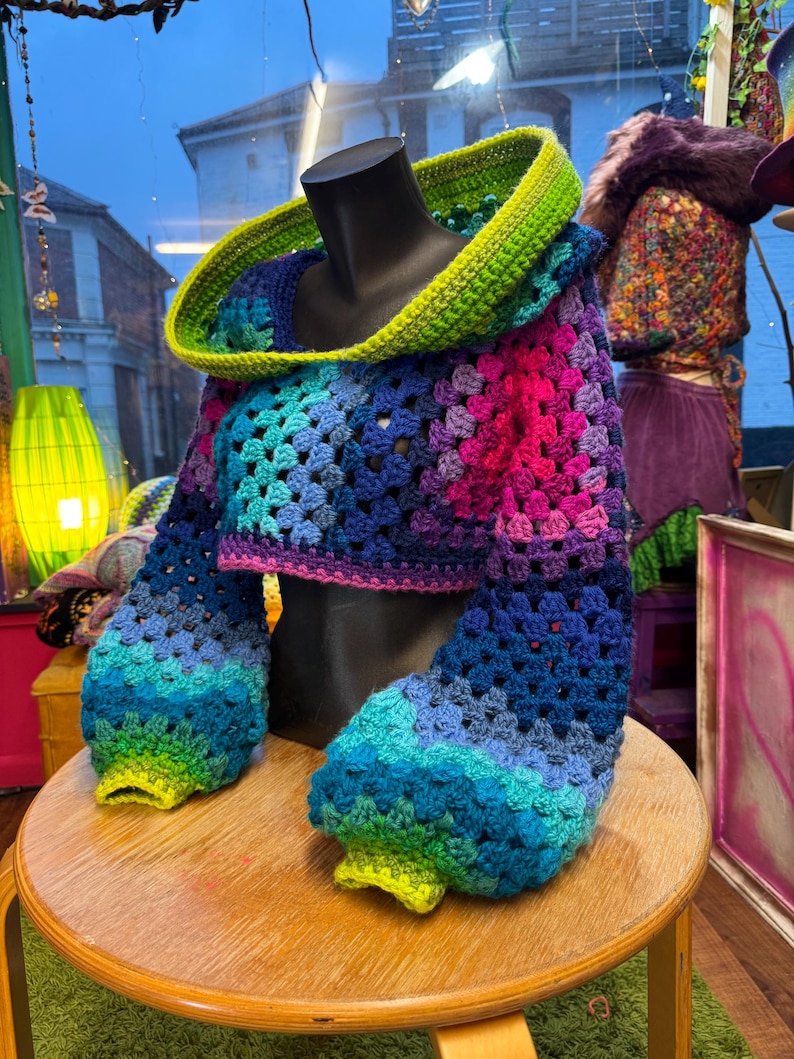 May include: A colorful, crocheted cropped sweater with a hood. The sweater features a vibrant gradient of blues, purples, and pinks, with a bright green hood and cuffs. The sleeves are long and puffy. The sweater is displayed on a black mannequin.