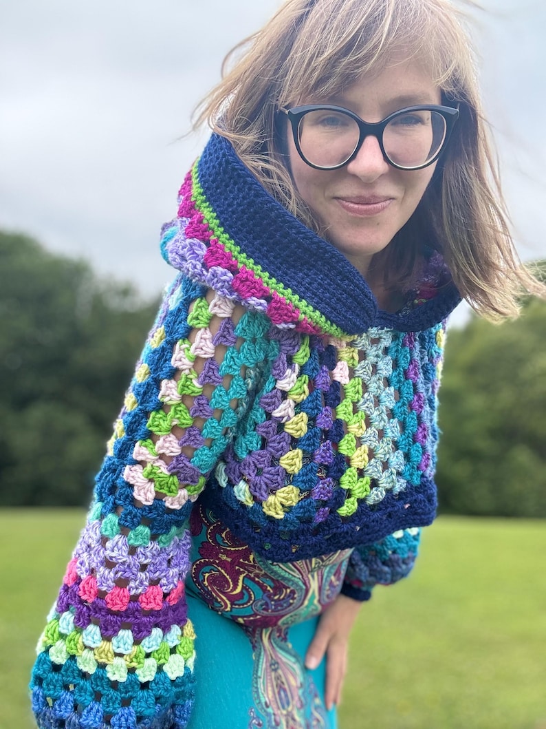 May include: A colorful, crocheted hooded sweater. The sweater features a granny square design with a mix of blue, purple, green, pink, and yellow yarn. The hood and cuffs are a solid dark blue. The sleeves are long and the sweater is cropped.