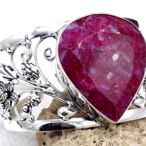 May include: A silver cuff bracelet featuring a large, faceted, teardrop-shaped ruby gemstone. The bracelet has an openwork design with floral and vine motifs. The ruby is a deep, rich red color, and the silver has a polished finish.