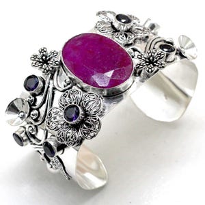 May include: A silver cuff bracelet with a large, faceted, oval magenta gemstone. The bracelet is decorated with floral and filigree patterns, and smaller purple gemstones. The design is intricate.