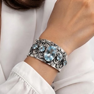 May include: A wide silver cuff bracelet featuring intricate scrollwork and floral metal detailing. The bracelet is set with five faceted light blue oval and round gemstones, with a large central oval stone as the primary focal point.