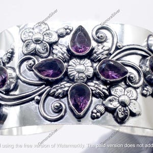 May include: A wide, silver cuff bracelet featuring floral and swirling patterns. The bracelet is embellished with multiple teardrop and round, deep purple gemstones. The cuff is approximately 5 cm wide.