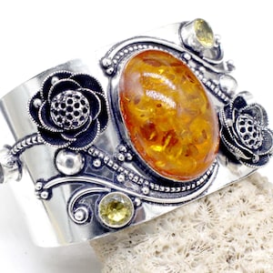 Op de afbeelding: Een brede zilveren manchetarmband met een ovale barnsteen als middelpunt. De armband is versierd met bloemen- en krulontwerpen en kleine gele edelstenen. De manchetarmband rust op een getextureerd, gebroken witte ondergrond.