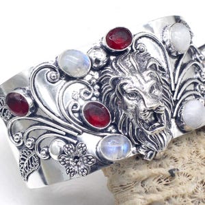 May include: A silver-toned cuff bracelet featuring a detailed lion's head design. The bracelet is adorned with red and white gemstones, and decorative floral and leaf accents. The cuff is resting on a textured, light-colored surface.