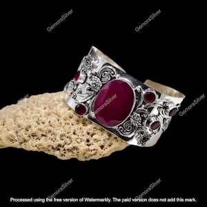 Könnte beinhalten: Ein breites silbernes Armreif-Armband mit einem ovalen, tiefroten Edelstein in der Mitte und kleineren roten Steinen und floralen Designs. Das Armband wird auf einer strukturierten, beigefarbenen Oberfläche präsentiert.