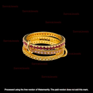 May include: A stack of three gold-colored rings. The top ring is adorned with red gemstones, the middle ring with pink gemstones, and the bottom ring with yellow gemstones. The rings are set against a black background.