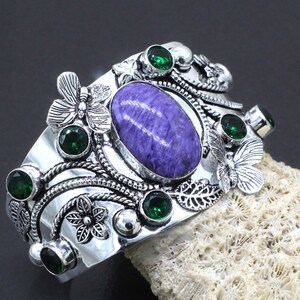 Pode incluir: Uma pulseira de punho prateada com uma grande pedra oval roxa no centro. A pulseira é adornada com pedras verdes, borboletas e detalhes florais. O design apresenta detalhes intrincados e um acabamento texturizado, criando uma joia única.