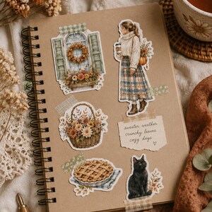 May include: A spiral-bound notebook with tan pages featuring various stickers including a window with a wreath, a person in a sweater and plaid skirt, a basket of flowers, a pie, a black cat, and a note with the text sweater weather crunchy leaves cozy days.