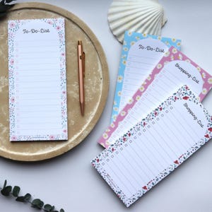 Könnte beinhalten: Eine Auswahl an Notizblöcken mit floralen Designs und einem Stift. Die Notizblöcke zeigen die Aufschriften "To-Do-List" und "Shopping-List" mit floralen Rändern in Rosa, Blau und Weiß. Ein roségoldener Stift liegt auf einem beigen Tablett.