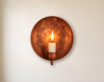 May include: A circular wall-mounted candle sconce with a textured, aged copper finish. The sconce features a wide, bowl-shaped backplate and a central cup holding a single white pillar candle with a bright, flickering yellow flame.