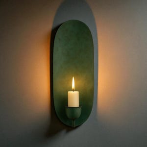 May include: A green wall sconce with a lit white candle. The sconce has an oval shape and a small holder for the candle. The background is a neutral gray wall, with a warm glow from the candle.
