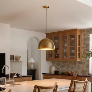 May include: A gold-coloured, dome-shaped pendant light hangs from the ceiling in a kitchen. The light fixture has a textured surface and illuminates the space below. The kitchen features wooden cabinets, a stone wall, and a white countertop.