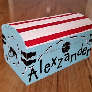 May include: A light blue wooden treasure chest with a red and white striped lid. The chest has a black and white design on the front and the name "Alexander" written in black letters.