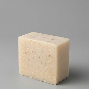 Puede incluir: Una pastilla de jabón rectangular de color beige claro, con motas visibles. El jabón tiene una textura ligeramente rugosa y se encuentra sobre un fondo gris liso. La pastilla es un bloque sólido, lo que sugiere un producto natural o artesanal.