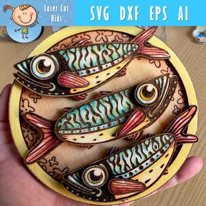 May include: Wooden artwork featuring three colorful fish arranged in a circular pattern. The fish have detailed designs with blue, red, and yellow accents. The artwork includes the text "SVG DXF EPS AI" and the logo "Laser Cut Kids".
