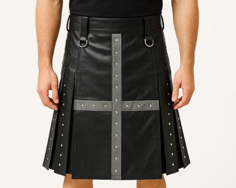 Op de afbeelding: Zwarte leren kilt met grijs kruisontwerp en zilveren klinknagels. De kilt heeft plooien en een riem met D-ringen. Het model draagt een zwart T-shirt en zwarte schoenen.