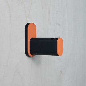 Puede incluir: Un gancho de pared moderno con un cuerpo cilíndrico negro y detalles en naranja. El gancho está montado sobre una placa trasera rectangular naranja. El diseño es minimalista y funcional.