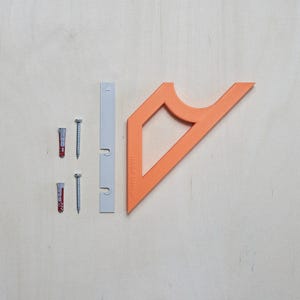 May include: An assortment of hardware components, including two screws, two wall anchors, a gray mounting bracket, and an orange angled bracket with the text "HAGA Design" printed on it. The items are arranged on a light wood surface.