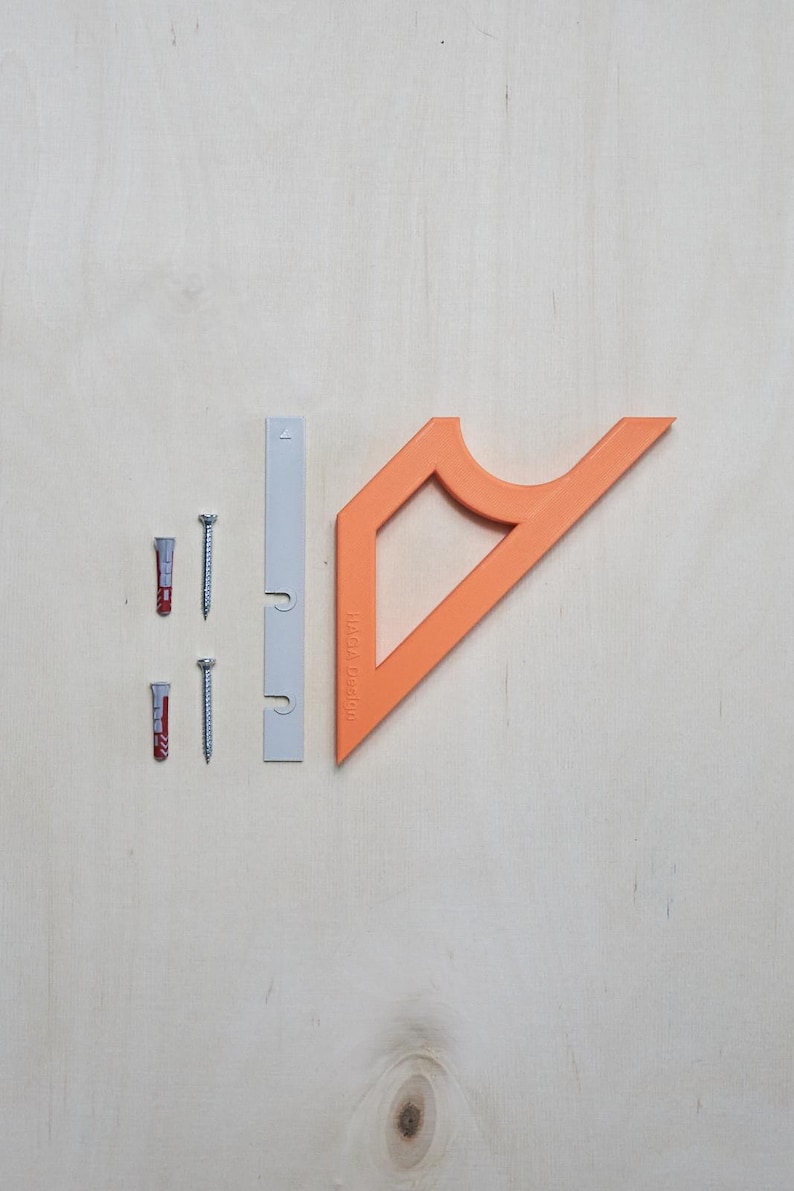 May include: An assortment of hardware components, including two screws, two wall anchors, a gray mounting bracket, and an orange angled bracket with the text "HAGA Design" printed on it. The items are arranged on a light wood surface.