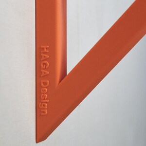 May include: Close-up of an orange geometric design element against a white background. The text "HAGA Design" is embossed on the side of the object. The design features angular shapes and a circular element.