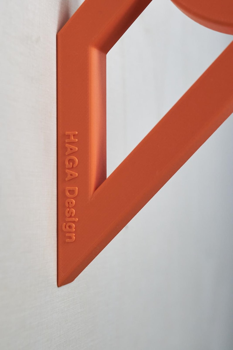 May include: Close-up of an orange geometric design element against a white background. The text "HAGA Design" is embossed on the side of the object. The design features angular shapes and a circular element.