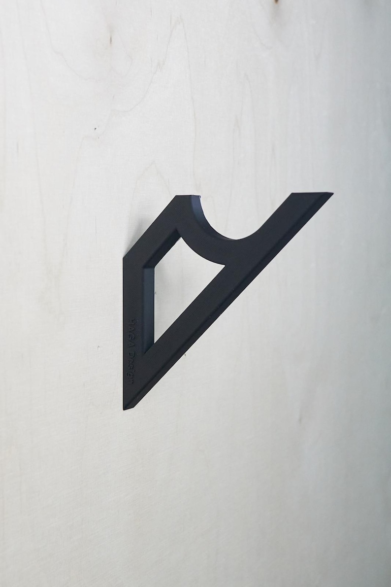 May include: A matte black wall-mounted coat hook with a geometric design. The hook features a triangular shape with a curved section and a diamond-shaped cutout. The text "Byga Design" is printed on the side.
