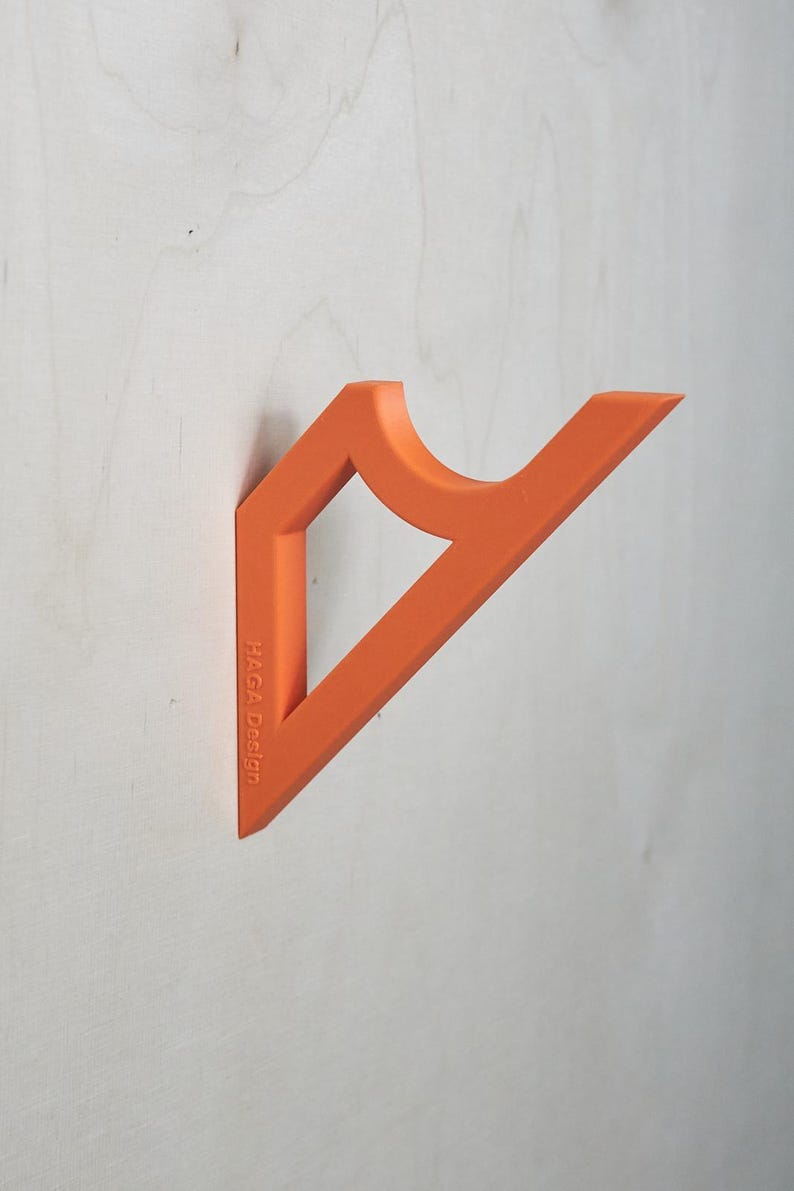 May include: Orange wall-mounted coat rack with a unique geometric design. The rack is made of a solid material and features a curved section for hanging items. The text "HAGA Design" is printed on the side.