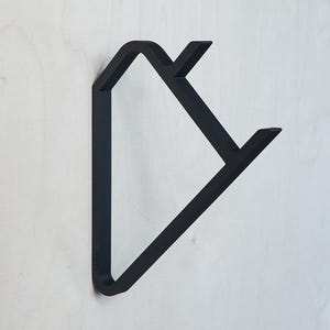 Puede incluir: Un gancho de pared minimalista de metal negro con un diseño geométrico. El gancho está montado sobre una superficie de madera de color claro. El diseño del gancho presenta una forma triangular con dos proyecciones anguladas.
